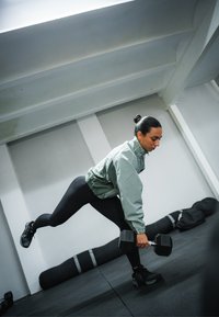 Idrottare i ljusgrön jacka och svarta träningstights håller i en svart hantel och utför ett utfall på gymgolvet med träningsmattor i närheten.