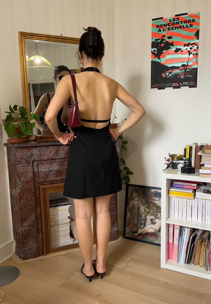 Femme portant une robe noire dos nu et des talons, tenant une pochette bordeaux, debout dans une pièce avec un miroir, une bibliothèque et une affiche au mur.