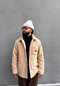 Μπεζ ζακέτα sherpa με κούμπωμα και τσέπη στο στήθος· συνδυασμένη με μαύρο τζόκερ και καρό καφέ παντελόνι. Γκρι πλεκτό σκουφί και γυαλιά ηλίου.