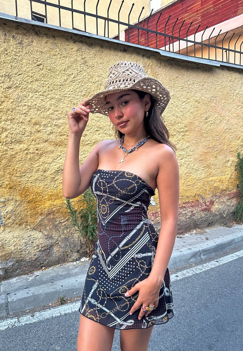 Jeune femme portant une robe sans bretelles à motifs et un chapeau tressé se tient dans une rue à côté d'un mur jaune texturé et d'une clôture en métal.
