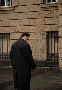 Mustast trentšmantlist, mis on lõdva lõikega, pikkade varrukatega ja nööbiga aksentidega, mille taustaks on tekstuurse kivimüür ja linnaline asfalt.