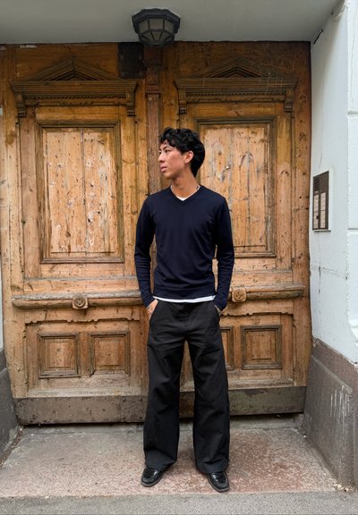 Joven con jersey azul marino y pantalones negros está de pie con las manos en los bolsillos, mirando hacia la izquierda, delante de unas grandes puertas de madera envejecida.