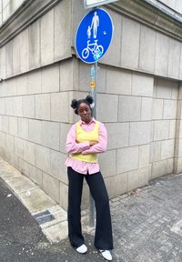 Cămașă în dungi roz și albă purtată peste un top galben, pantaloni negri largi, adidași albi, stând lângă un semn albastru pentru pietoni și bicicliști.