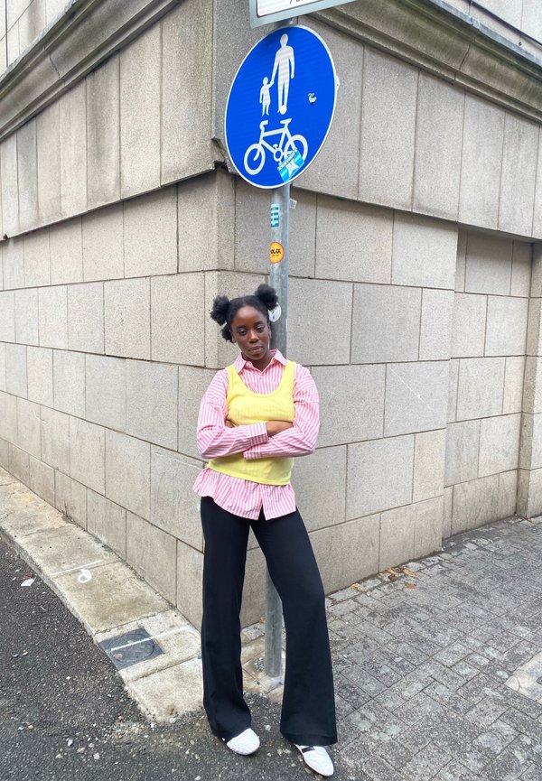 Prugasta ružičasto-bijela košulja preko žute majice, crne široke hlače, bijele tenisice, stoji pored plave oznake za pješake i bicikliste.