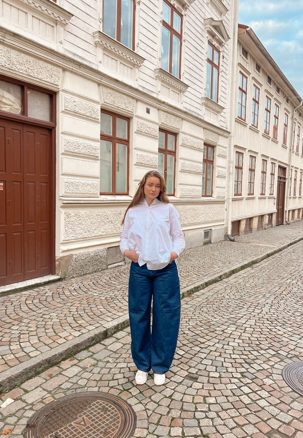 White button-up shirt, loose denim wide-leg pants, and white sneakers, standing on a cobblestone street beside historic brick buildings.