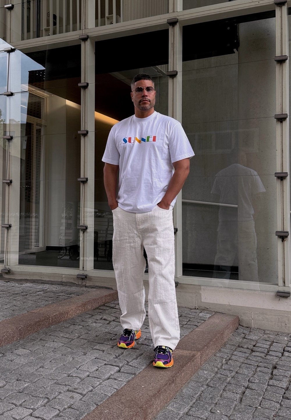 White short-sleeved t-shirt featuring colourful "SUNSET" text, worn with loose white textured trousers and multicoloured sports shoes.