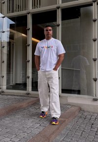 White short-sleeved t-shirt featuring colourful "SUNSET" text, worn with loose white textured trousers and multicoloured sports shoes.