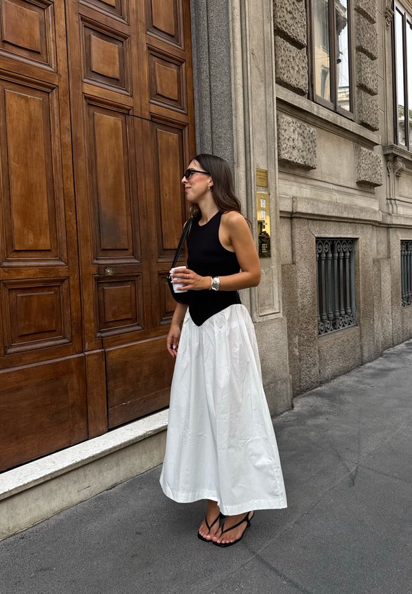 Femme en haut noir sans manches et jupe blanche tenant une tasse, debout près de grandes portes en bois dans une rue de la ville.
