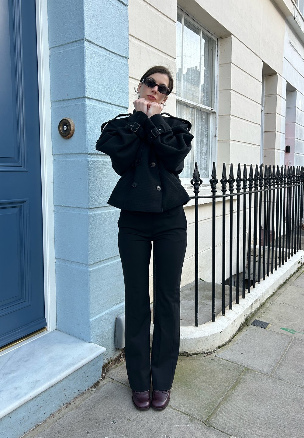 Veste noire à double boutonnage avec manches surdimensionnées, pantalon noir ajusté et lunettes de soleil foncées. Chaussures bordeaux foncé sur un pavé en pierre.