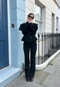 Veste noire à double boutonnage avec manches surdimensionnées, pantalon noir ajusté et lunettes de soleil foncées. Chaussures bordeaux foncé sur un pavé en pierre.