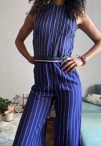 Γυναίκα με ολόσωμη φόρμα μπλε και λευκές κάθετες ρίγες, με τα χέρια στη μέση, φορώντας βραχιόλια από κόκκινα και κίτρινα χάντρες, που στέκεται σε εσωτερικό χώρο κοντά σε ένα τραπέζι.