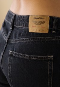 Tumši džinsi no denima ar ādas etiķeti aizmugurē, kurā iekļauti kopšanas norādījumi. Izmēra etiķete norāda 26. Redzamas kabatas un šuvju detaļas.