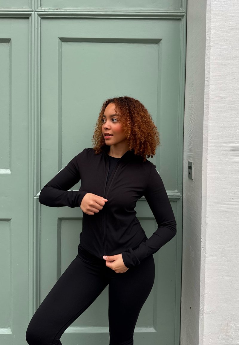 Veste zippée noire à manches longues en tissu extensible, associée à des leggings noirs. En arrière-plan, une porte vert clair à panneaux.