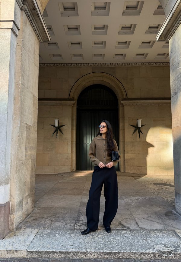Una mujer con gafas de sol, una chaqueta marrón y pantalones negros está de pie bajo un arco de piedra, frente a una gran entrada con verja.