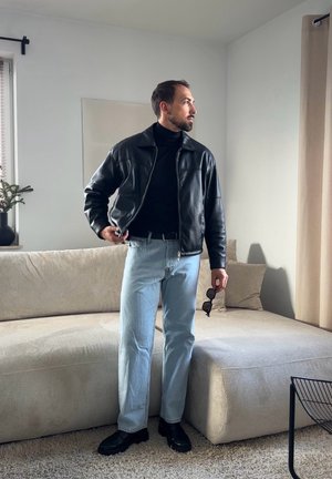 Hombre con chaqueta de cuero negra y jeans azul claro sostiene unas gafas de sol mientras está de pie en una sala de estar con sofá beige y cortinas.