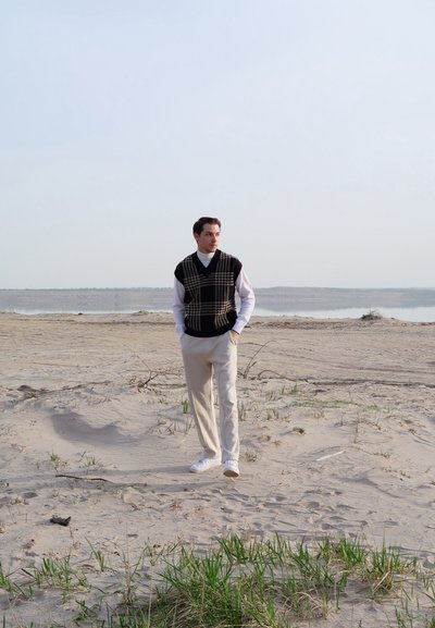 Suéter sin mangas de cuadros negros y beige sobre un cuello de tortuga blanco, combinado con pantalones beiges claros y zapatillas blancas, en una playa de arena.