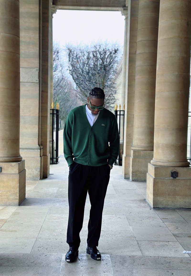 Mann in grünem Strickjacke und schwarzer Hose steht mit den Händen in den Taschen unter einem Steinbogen und schaut nach unten.