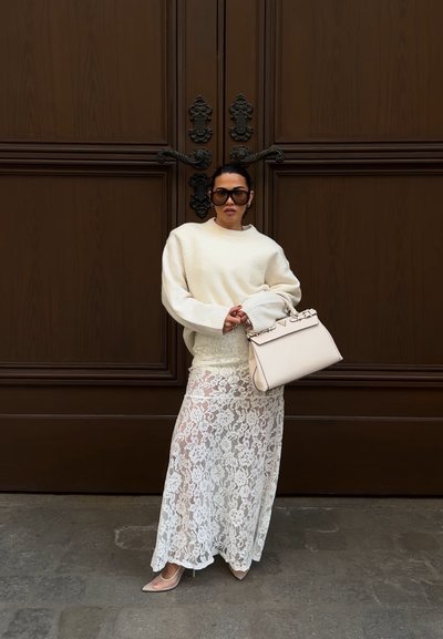 Mujer con suéter crema de gran tamaño, falda blanca de encaje, tacones beige, gafas de sol grandes, sosteniendo bolso crema, de pie frente a grandes puertas marrones ornamentadas.