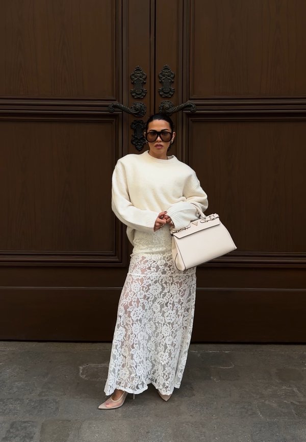 Žena nosiaca nadmerný krémový sveter, bielu čipkovanú sukňu, béžové podpätky, veľké slnečné okuliare, drží krémovú kabelku, stojí pri veľkých zdobených hnedých dverách.