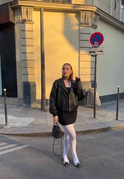 Chaqueta bomber de cuero negra, vestido mini negro, medias blancas con estampado, bolso negro y zapatos planos negros, con una pared amarilla y señales de tráfico en el fondo.