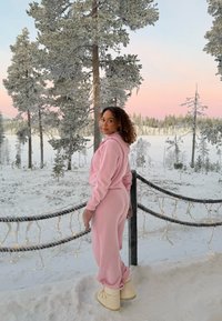 Vrouw in roze loungewear en beige laarzen staande op een besneeuwd balkon met bevroren bomen en een pastelkleurige zonsondergang aan de hemel op de achtergrond.