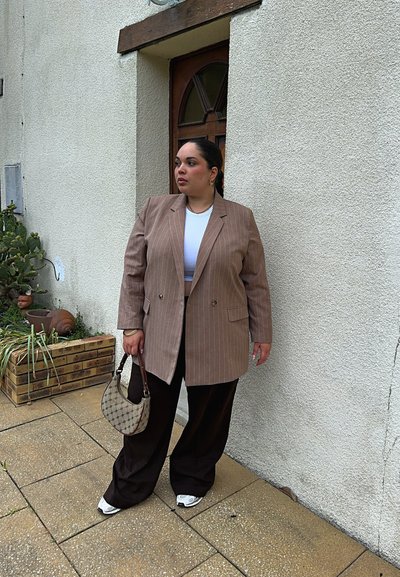 Blazer ancho color marrón con rayas sobre una blusa blanca, combinado con pantalones anchos negros y zapatillas blancas. Llevando un bolso de estampado.