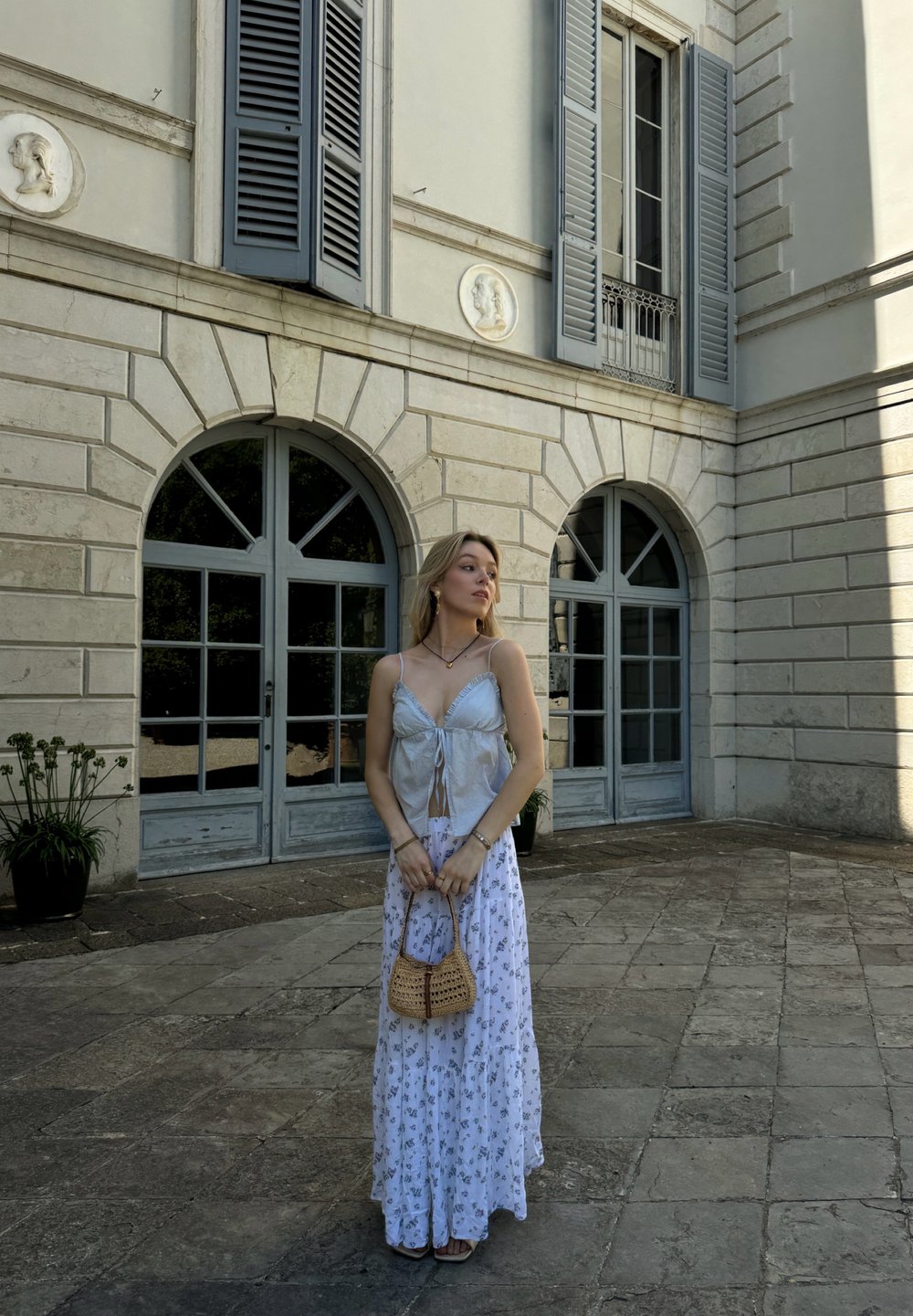 Top de seda azul claro com alças finas e frente atada, combinado com uma saia longa branca com padrões florais; segurando uma bolsa trançada.
