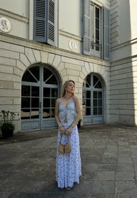 Top de seda azul claro com alças finas e frente atada, combinado com uma saia longa branca com padrões florais; segurando uma bolsa trançada.