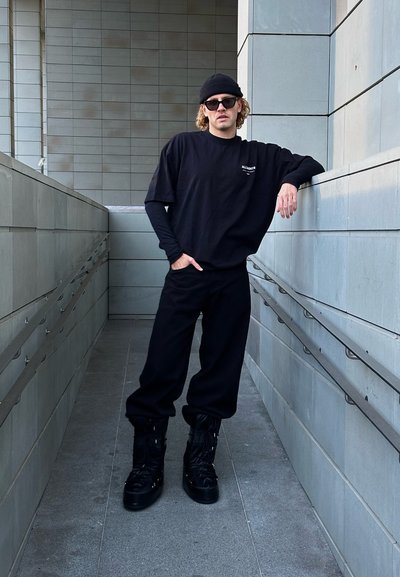 Camiseta negra de gran tamaño con logo, pantalones deportivos negros a juego y botas negras gruesas. El modelo se apoya contra una pared gris en un pasillo moderno.