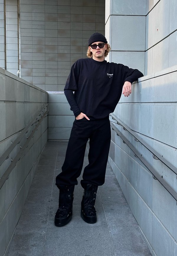 Oversized sort t-shirt med logo, matchende sorte joggers og kraftige sorte støvler. Modellen læner sig op ad en grå væg i en moderne korridor.