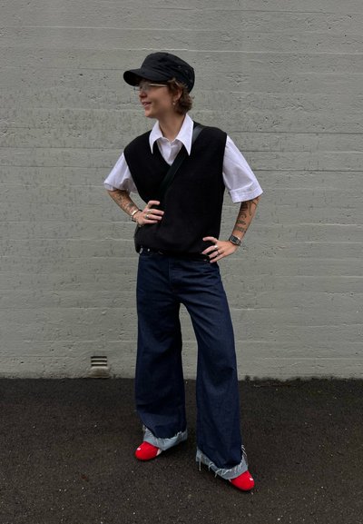 Chaleco de punto negro sobre una camisa blanca con cuello, combinado con jeans de mezclilla oscuros de pierna ancha y zapatos de punta roja con puños desgastados.