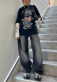 Svart grafisk t-skjorte med et design av hodeskalle og hjerte. Kombinert med vide, falmede grå jeans. Hvite langermede topper synlige under.