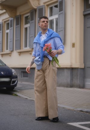 Joven con pantalones beige de pierna ancha y camisa azul con suéter sobre los hombros, sosteniendo un ramo de flores rosas en una calle de la ciudad.
