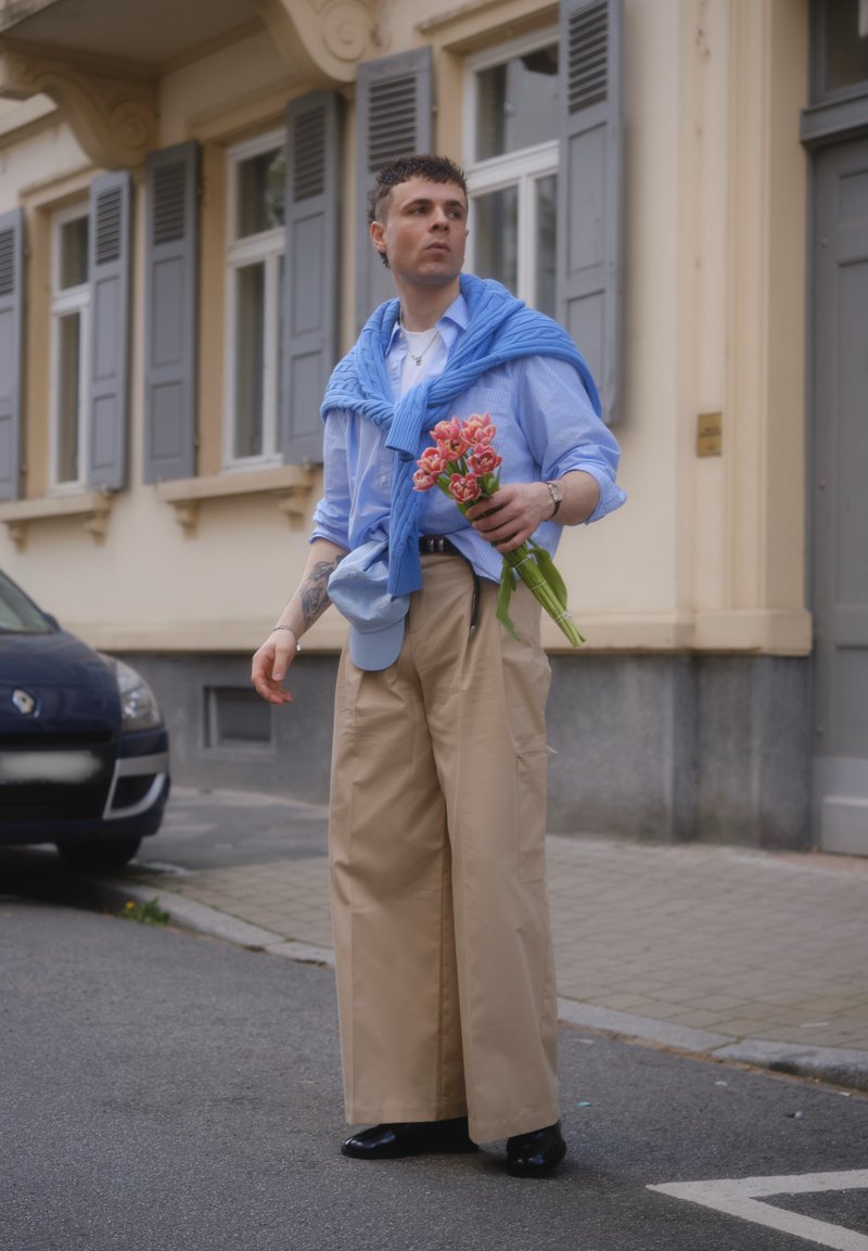 Jeune homme portant un pantalon large beige et une chemise bleue avec un pull posé sur les épaules, tenant un bouquet de fleurs roses dans une rue de la ville.