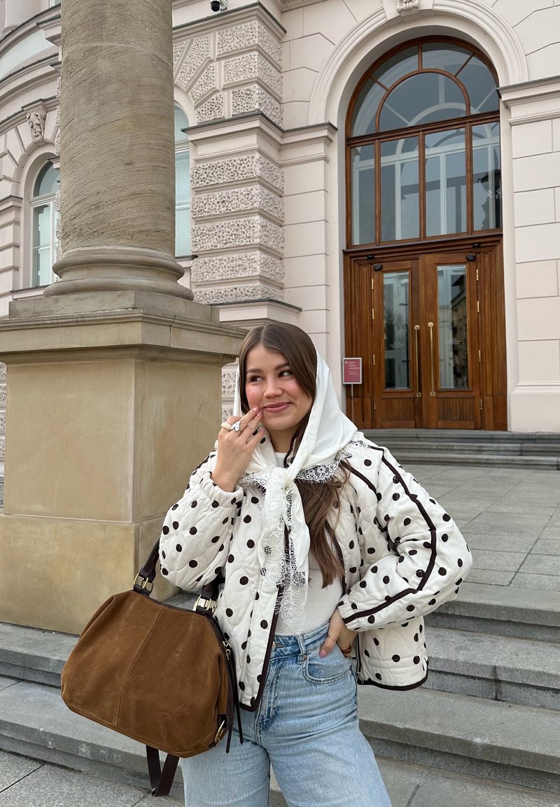 Jeune femme en veste à pois et foulard blanc sur la tête, debout près d'une colonne en pierre à l'extérieur d'un bâtiment avec des portes doubles en bois. Sac marron au bras.