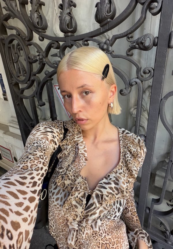 Femme aux cheveux blonds courts avec une pince noire porte un haut à volants imprimé léopard, prenant un selfie devant une porte en fer noir ornée.