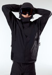 Άτομο που φοράει μαύρο μπουφάν με κουκούλα και παντελόνι, με σκούρα γυαλιά σκι, ρυθμίζοντας την κουκούλα με τα χέρια πίσω από το κεφάλι, σε απλό φωτεινό φόντο.