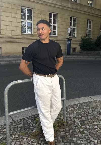 Camiseta negra de manga corta combinada con pantalones de lino blancos. La persona se apoya en una barandilla de metal gris en una calle de adoquines.