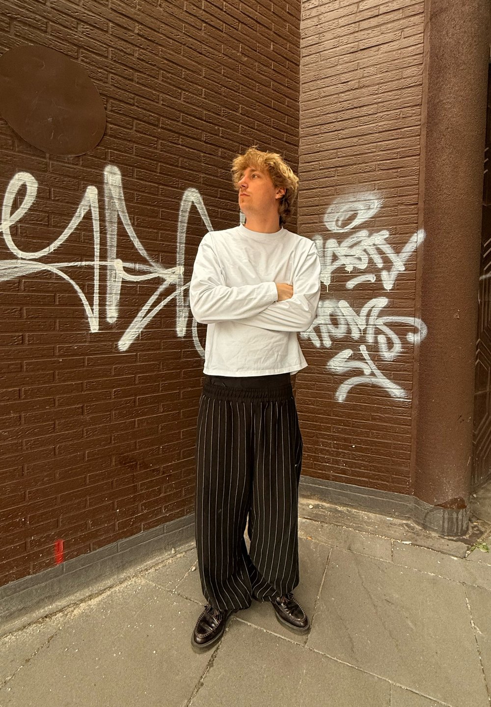 Hvid langærmet skjorte, sorte stribede vide bukser og sorte sko, står mod en tekstureret brun væg med graffiti.