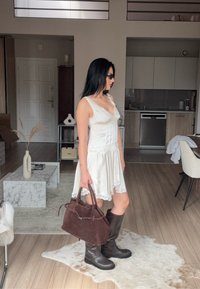 Vrouw in een witte kanten jurk, met donkere kniehoge laarzen en een zonnebril, die een bruin suède handtas vasthoudt in een moderne woonkamer.