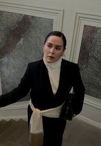 Sort skræddersyet blazer over en hvid højhalset trøje, spændt med et hvidt frynsesjal, holder en sort håndtaske, på en trappe med dekorative vægge.