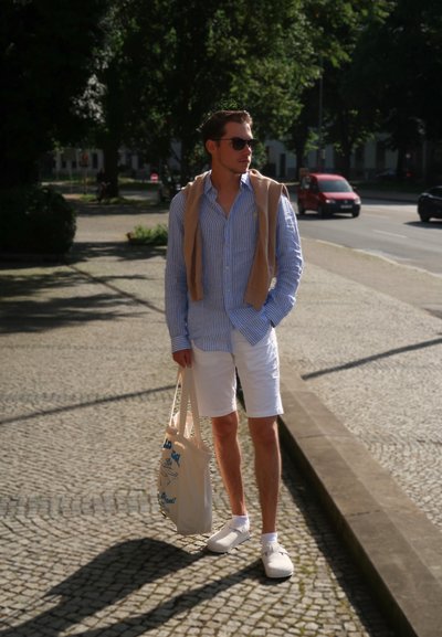 Camisa de botones a rayas azul claro, pantalones cortos blancos, zapatos crema, suéter beige colgado sobre los hombros, bolsa tote beige con texto azul.
