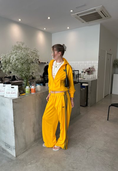 Chándal amarillo en una tela suave, con rayas laterales negras y chaqueta con cremallera. Combinado con sandalias blancas y accesorios dorados en capas.
