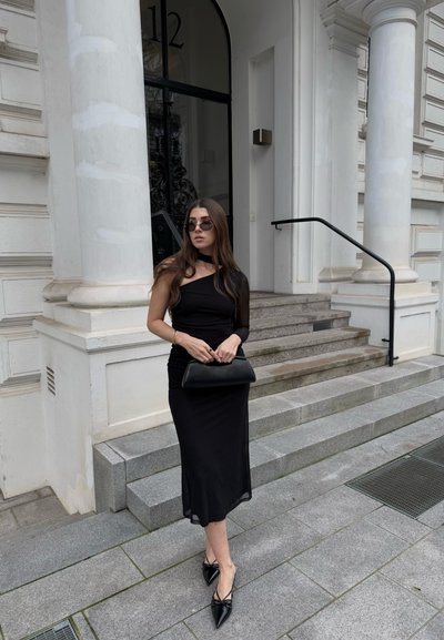 Vestido negro ajustado con un diseño de un solo hombro, combinado con bailarinas negras de punta y un clutch a juego. Tela suave, textura mínima.