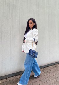 Blazer blanco sobre una blusa blanca, combinado con jeans de pierna ancha azul y zapatillas blancas. Sosteniendo un bolso acolchado azul oscuro con una correa de cadena.