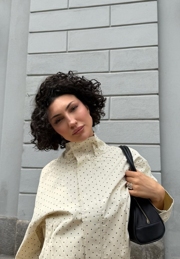Femme aux cheveux courts et bouclés porte une veste claire à pois et tient un sac à main noir sur son épaule devant un mur de briques gris.