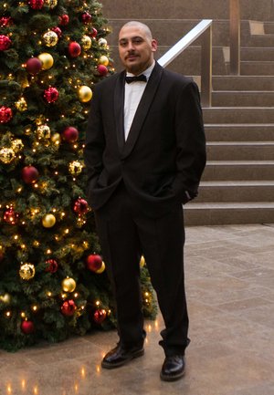 Esmoquin negro con pajarita, de pie frente a un árbol de Navidad decorado con adornos rojos, dorados y luces parpadeantes.