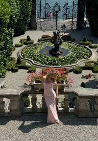 Rosa pallido abito in pizzo con silhouette aderente e design off-the-shoulder. Ambientazione da giardino con una fontana e fiori in fiore sullo sfondo.