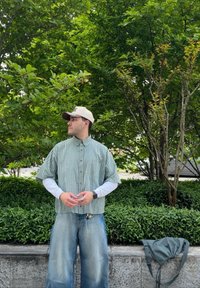 Grönrutigt kortärmat skjort över en vit långärmad skjort, ihop med ljusblå baggy jeans. En beige keps och en grön väska är synliga.