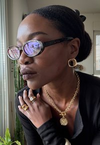 Owalne okulary przeciwsłoneczne z lustrzanymi fioletowymi soczewkami, złota naszyjnik z zawieszką, masywne złote pierścionki i kwadratowe kolczyki na tle ciemnej, prążkowanej bluzki.